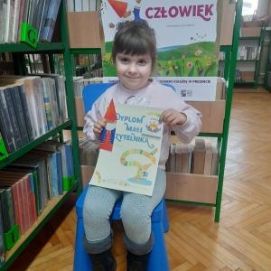 W pomieszczeniu biblioteki. Uśmiechnięta dziewczynka siedzi na niebieskim krześle. W ręku trzyma dyplom. W tle regały z książkami.