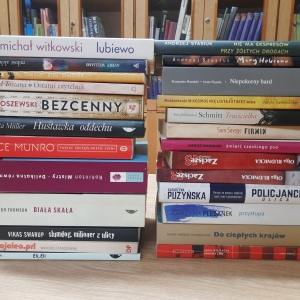W bibliotece. Na stoliku ułożone dwa stosy książek. W tle regały z książkami.