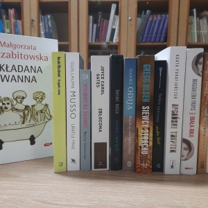 W bibliotece. Na stoliku ustawione książki. W tle regały z książkami.