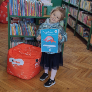 Wnętrze biblioteki. Dziewczynka stoi obok pomarańczowej pufy, w ręku trzyma dyplom w kolorze niebieskim, na którym jest grafika delfina w kolorze różowym. W tle widać zielone regały z książkami