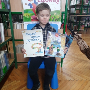 W bibliotece. Chłopiec siedzi na niebieskim krześle. W ręku trzyma książkę i dyplom.