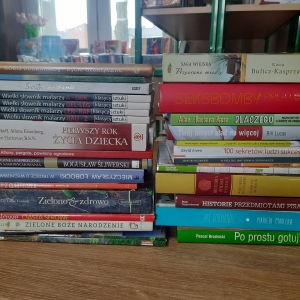 W bibliotece. Na stoliku ustawione książki. W tle regały z książkami.