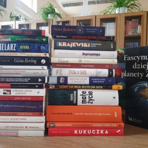 W bibliotece. Na stoliku ustawione książki. W tle regały z książkami.