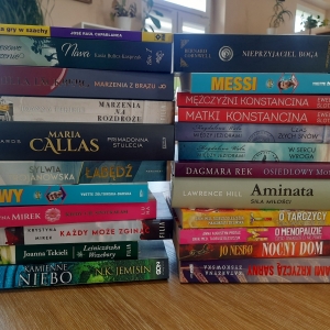 W bibliotece. Na stoliku ustawione książki. W tle regały z książkami.