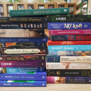 W bibliotece. Na stoliku ustawione książki. W tle regały z książkami.
