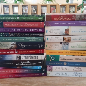 W bibliotece. Na stoliku ustawione książki. W tle regały z książkami.