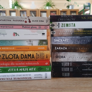W bibliotece. Na stoliku ustawione książki. W tle regały z książkami.