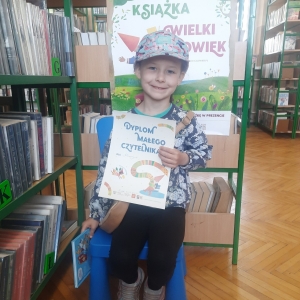 W pomieszczeniu biblioteki. Uśmiechnięta dziewczynka siedzi na niebieskim krześle. W ręku trzyma dyplom. W tle regały z książkami.
