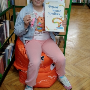 W pomieszczeniu biblioteki. Uśmiechnięta dziewczynka siedzi na pomarańczowej pufie. W ręku trzyma dyplom. W tle regały z książkami.