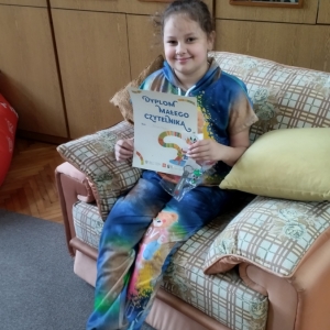 W pomieszczeniu biblioteki. Uśmiechnięta dziewczynka siedzi na beżowym fotelu. W ręku trzyma dyplom. W tle regały z książkami.
