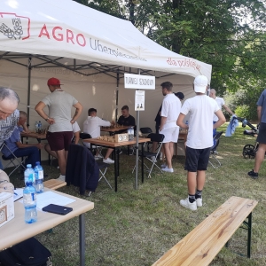 W plenerze. W trakcie trwania turnieju szachowego. Przy stolikach siedzą gracze, obok stoją osoby obserwujące.