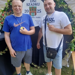W plenerze. Dwóch mężczyzn stoi przy ściance dekoracyjnej i pokazują otrzymane medale.