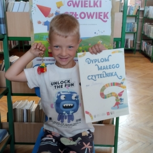 W bibliotece. Chłopiec siedzi na niebieskim krześle. W ręku trzyma dyplom. W tle regały z książkami.