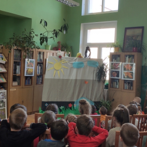 W pomieszczeniu. Grupa dzieci ogląda teatrzyk. Nad kotarą widać 3 maskotki. W tle regały z książkami