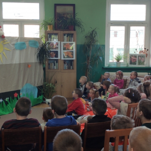 W pomieszczeniu. Grupa dzieci ogląda teatrzyk. Nad kotarą widać  maskotkę - kaczuszkę. Obok sceny widać panią bibliotekarkę. W tle regały z książkami