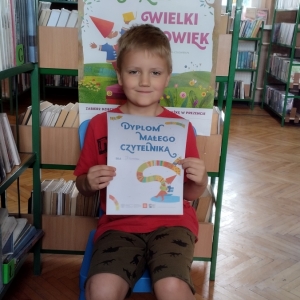 W bibliotece. Uśmiechnięty chłopiec siedzi na niebieskim krześle. W ręku trzyma dyplom. W tle regały z książkami.