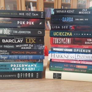 W bibliotece. Na stoliku ustawione książki. W tle regały z książkami.