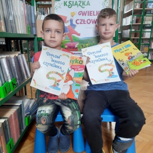  	W bibliotece. Dwaj chłopcy siedzą na niebieskich krzesłach. Każdy trzyma dyplom i książkę.
