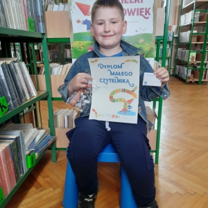 W bibliotece. Uśmiechnięty chłopiec siedzi na niebieskim krześle. W ręku trzyma dyplom. W tle regały z książkami.