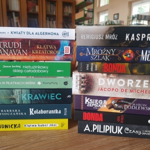 W bibliotece. Na stoliku ustawione książki. W tle regały z książkami.
