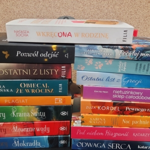 W bibliotece. Na stoliku ustawione książki. W tle regały z książkami.