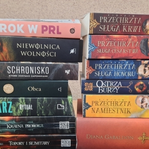 W bibliotece. Na stoliku ustawione książki. W tle regały z książkami.
