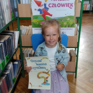 W pomieszczeniu biblioteki. Uśmiechnięta dziewczynka siedzi na niebieskim krześle. W ręku trzyma dyplom. W tle regały z książkami.