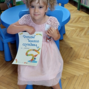 W pomieszczeniu biblioteki. Uśmiechnięta dziewczynka siedzi na niebieskim krześle. W ręku trzyma dyplom. W tle regały z książkami.