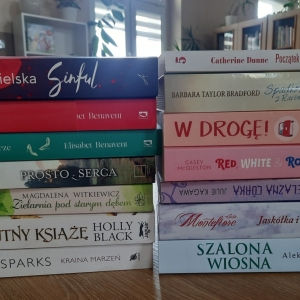 W bibliotece. Na stoliku ustawione książki. W tle regały z książkami.