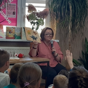 W bibliotece. Grupa osób patrzy na prowadzącą spotkanie. Autorka pokazuje swoją książkę.