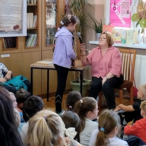 W bibliotece. Kobieta siedzi przy stoliku i trzyma w ręku mikrofon. Obok niej stoi dziewczynka i odpowiada na zadane pytanie.
