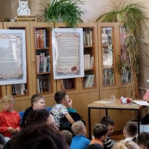 W bibliotece. Grupa dzieci patrzy na prowadzącą spotkanie. Kobieta siedzi przy stoliku, w ręku trzyma mikrofon.