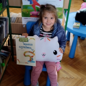 W bibliotece. Uśmiechnięta dziewczynka siedzi na niebieskim krześle. W ręku trzyma dyplom i pluszową poduszkę w kształcie kota. W tle regały z książkami.