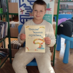 W bibliotece. Uśmiechnięty chłopiec siedzi na niebieskim krześle. W ręku trzyma dyplom. W tle regały z książkami.