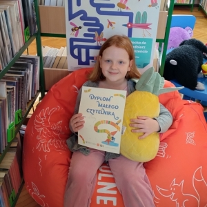 W bibliotece. Uśmiechnięta dziewczynka siedzi na pomarańczowej pufie. W ręku trzyma dyplom i pluszową poduszkę w kształcie ananasa. W tle regały z książkami.