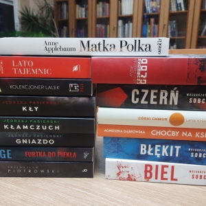 W bibliotece. Na stoliku ustawione książki. W tle regały z książkami.