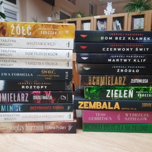 W bibliotece. Na stoliku ustawione książki. W tle regały z książkami.