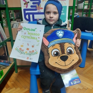 W bibliotece. Chłopiec siedzi na niebieskim krześle. W ręku trzyma dyplom i maskotkę. W tle regały z książkami.