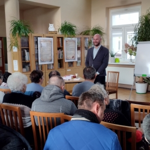W bibliotece. Grupa osób patrzy na mężczyznę, który prowadzi spotkanie.