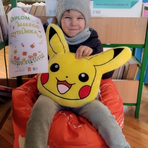 W bibliotece. Uśmiechnięty chłopiec siedzi na pomarańczowej pufie. W ręku trzyma dyplom i pluszową poduszkę w kolorze żółtym. W tle regały z książkami.