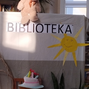 W bibliotece. Scenografia przygotowana na potrzeby teatrzyku dla dzieci. Nad kotarą pluszowy miś. Na kotarze napis "Biblioteka". Poniżej na krześle siedzi maskotka - pluszowy miś z koszyczkiem.