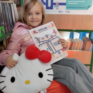 W bibliotece. Uśmiechnięta dziewczynka siedzi na pomarańczowej pufie. W ręku trzyma dyplom i pluszową poduszkę w kształcie kota. W tle regały z książkami.