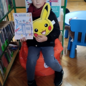 W bibliotece. Chłopiec siedzi na pomarańczowej pufie. W ręku trzyma dyplom i pluszową poduszkę w kolorze żółtym. W tle regały z książkami.