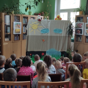 W budynku. Grupa przedszkolaków ogląda teatrzyk. Dzieci zwrócone są w kierunku sceny przedstawiającej łąkę ze stawem. Nad sceną dwie pluszowe kaczuszki, biała i żółta. W tle szafy i regały z książkami, okna oraz kwiaty doniczkowe.