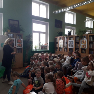 W budynku. Grupa małych dzieci siedzi na krzesłach i patrzy w kierunku stojącej kobiety. Kobieta mówi do dzieci. Za kobietą dekoracje przedstawiające łąkę ze stawem. W tle szafy i regały z książkami.