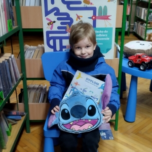 W bibliotece. Uśmiechnięty chłopiec siedzi na niebieskim krześle. W ręku trzyma dyplom i pluszową poduszkę w kolorze niebieskim. W tle regały z książkami.