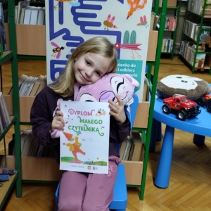 W bibliotece. Uśmiechnięta dziewczynka siedzi na niebieskim krześle. W ręku trzyma dyplom i pluszową poduszkę w kolorze różowym. W tle regały z książkami.