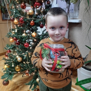 W bibliotece. Uśmiechnięty chłopiec stoi przy choince, trzyma słoik z cukierkami.