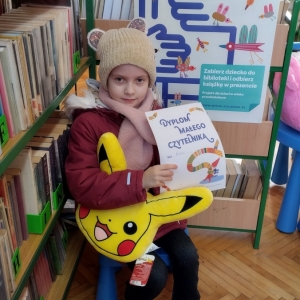 W bibliotece. Uśmiechnięta dziewczynka siedzi na niebieskim krześle. W ręku trzyma dyplom i pluszową poduszkę w kolorze żółtym. W tle regały z książkami.