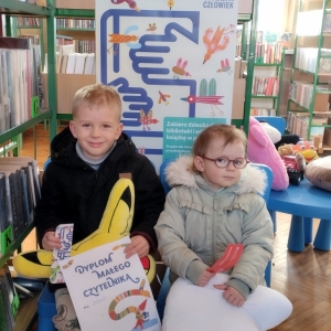 W bibliotece. Dwoje dzieci siedzi na niebieskich krzesłach. W tle regały z książkami.